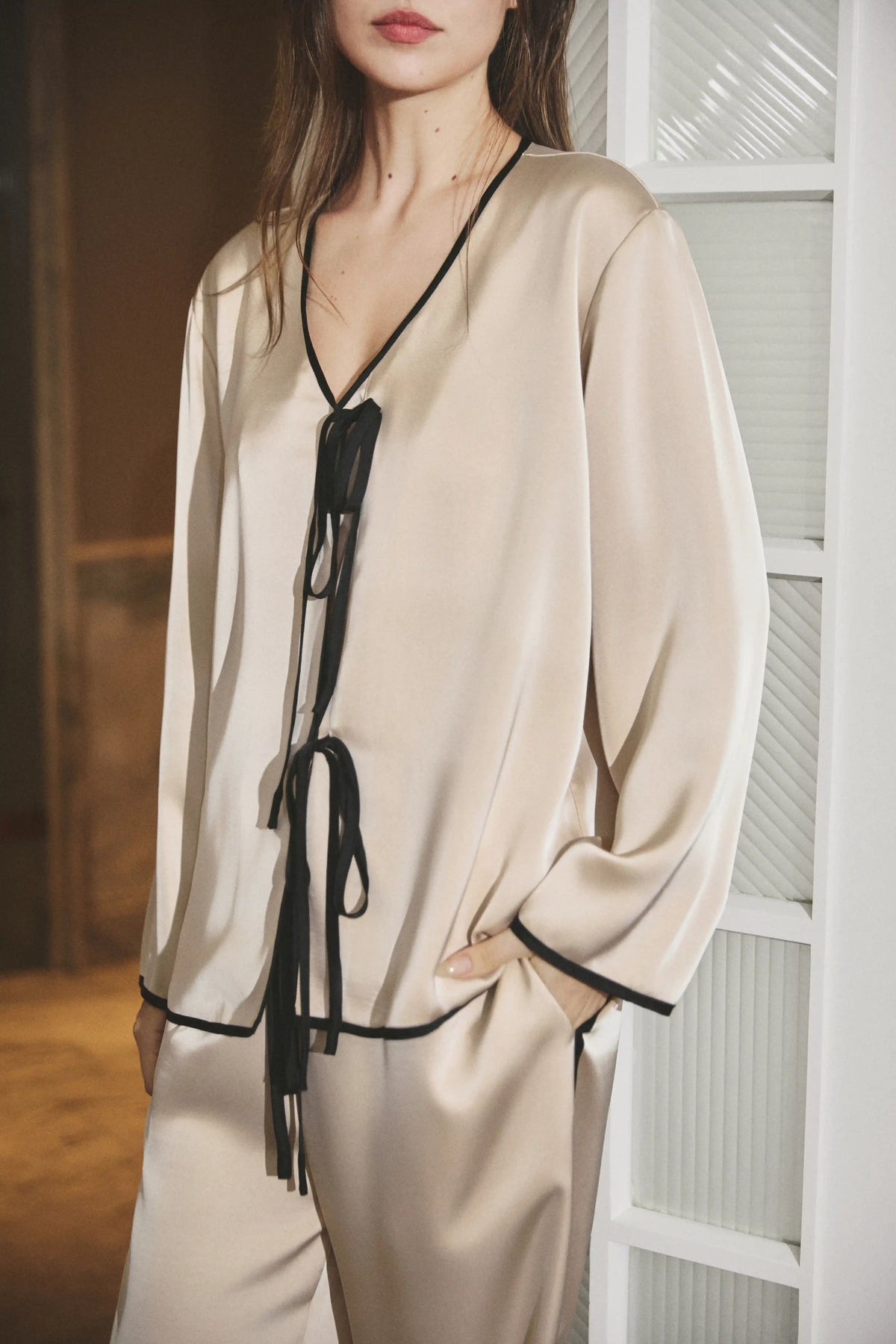 A woman wearing a beige satin pajama set with black trim and tie details, standing indoors next to a white paneled door.