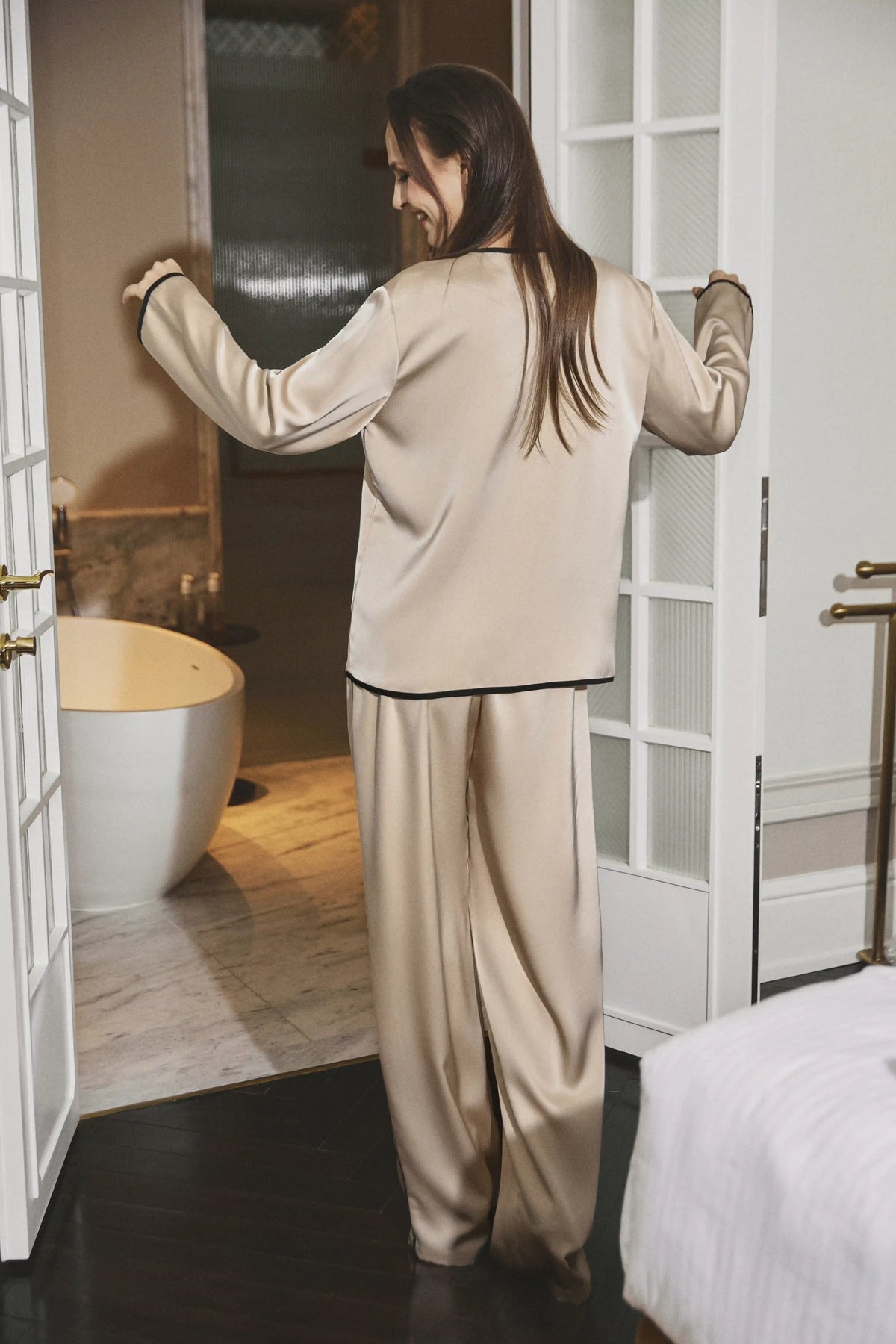 A woman wearing a beige satin pajama set with black piping trim, standing in a bedroom doorway next to a bathtub.
