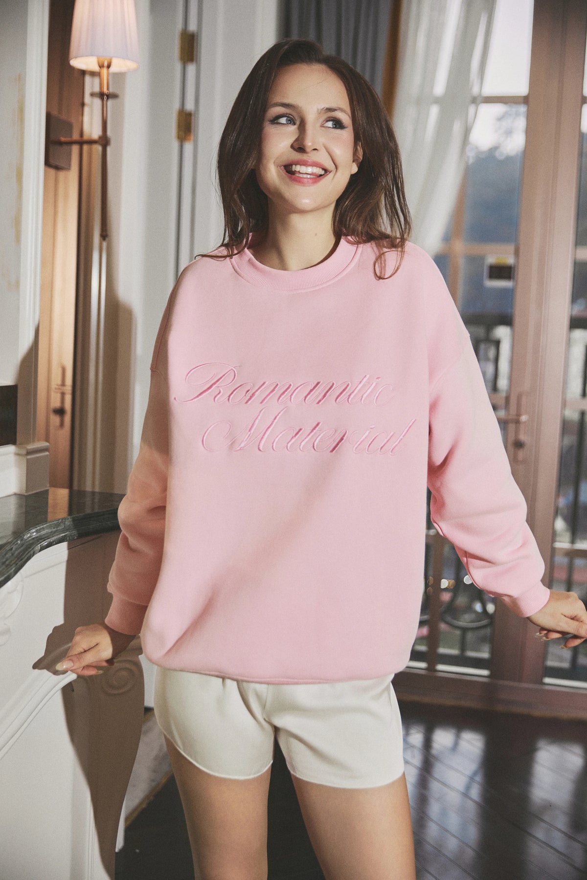 A woman wearing a light pink crewneck sweatshirt with 'Romantic Material' embroidered in pink script across the chest, paired with off-white shorts, standing in a warmly lit indoor setting.