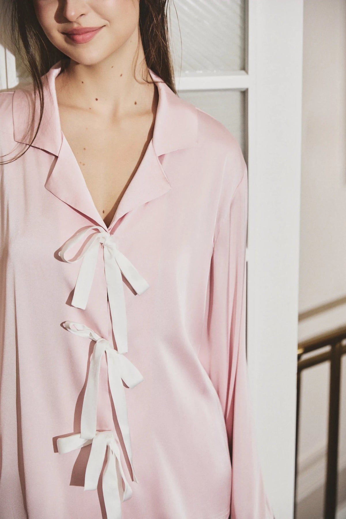 A close-up of a woman wearing a light pink long-sleeve pajama shirt with white tie-front ties, shown in a softly lit indoor setting.