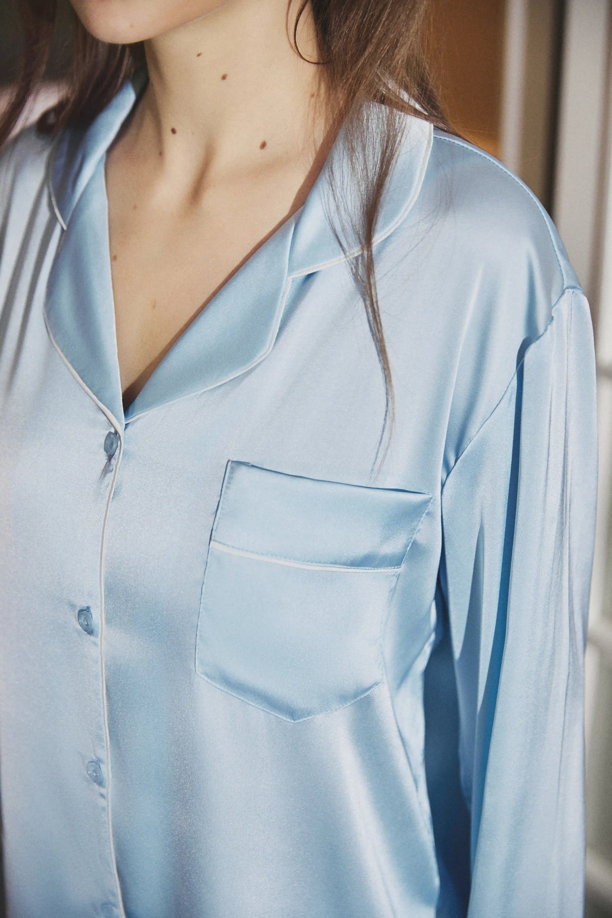 A close-up of a person wearing a light blue satin pajama shirt with a collar, button front, and chest pocket.
