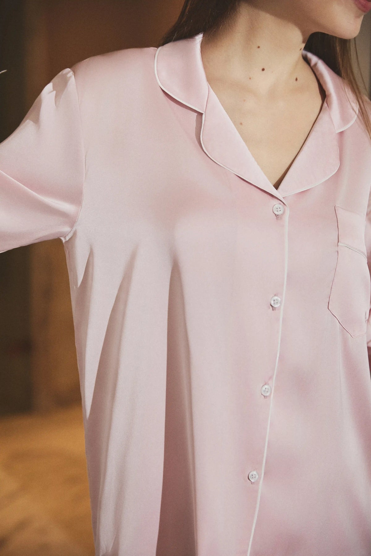 A close-up of a person wearing a light pink satin pajama shirt with a collar, button front, and chest pocket, photographed indoors against a softly blurred warm brown background.