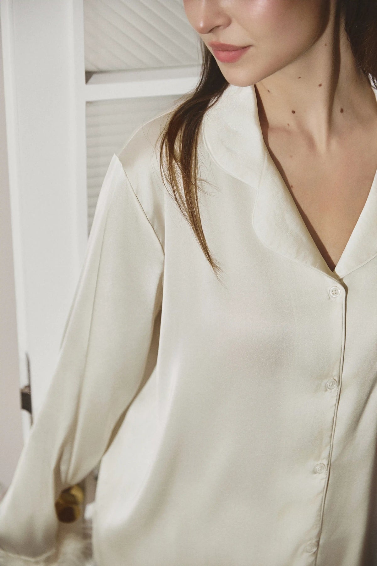 A close-up of a woman wearing a cream-colored silk button-up shirt. The image is cropped to show her neck, lower face, and the shirt, with a soft, warm-toned background.