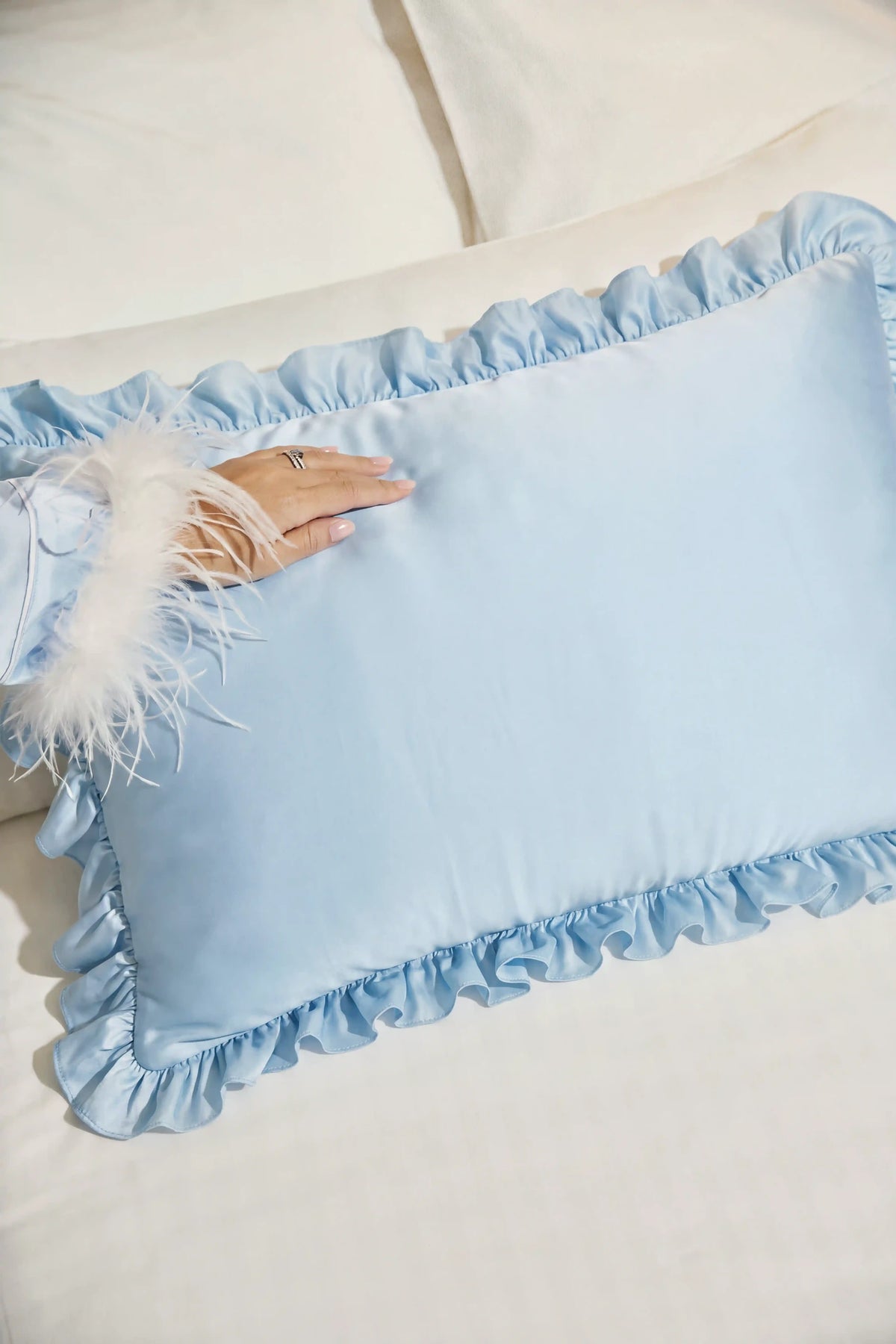 A light blue satin pillow with ruffled trim and a white feather accent, resting on a white bed with a hand touching the satin surface.